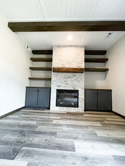 White brick fireplace with floating wood shelves, dark beams, and engineered hardwood flooring