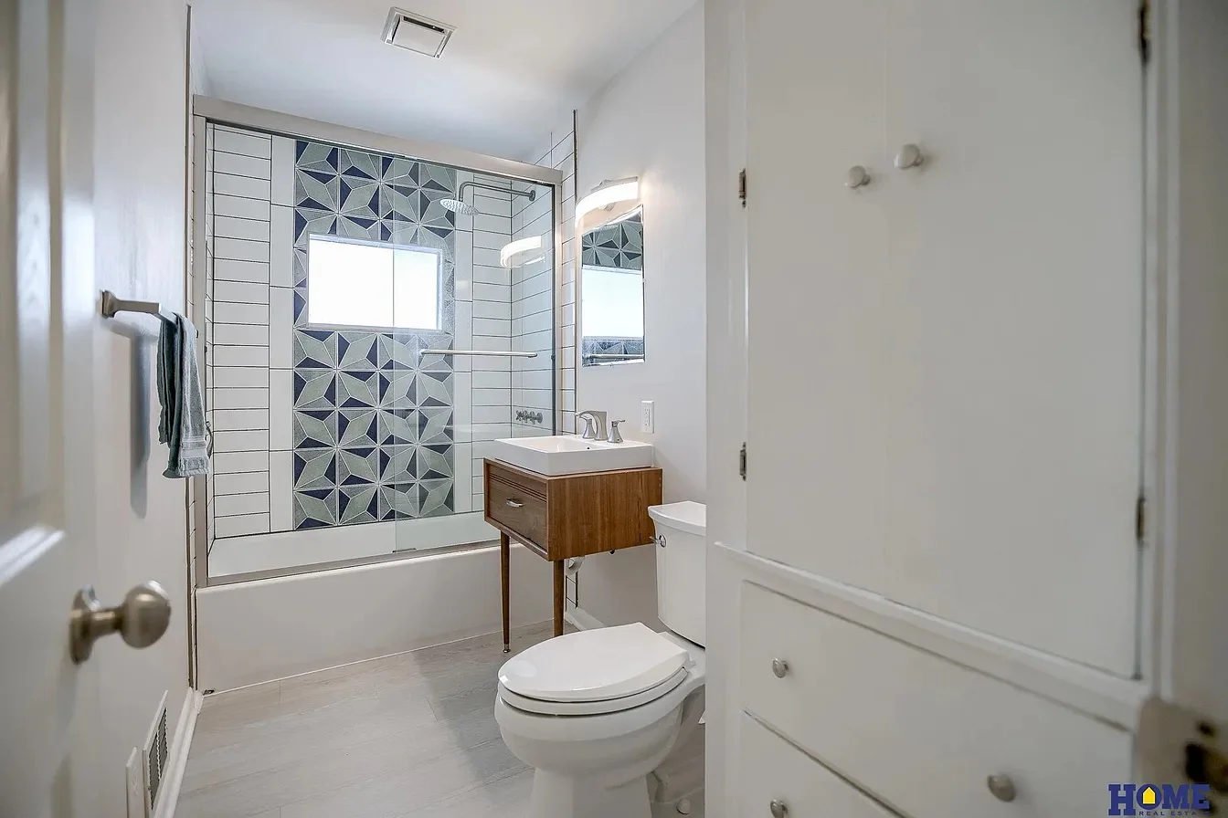 Bathroom with geometric tile shower, white subway tiles, frameless glass door, wood vanity, and white cabinetry