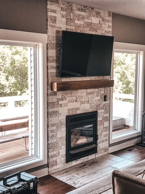 Stacked stone fireplace with black insert, wood mantel, and mounted TV between glass doors